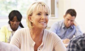 A senior woman smiling and sitting in class, looking up from her paper.