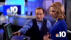 A middle aged man points at a computer screen while a young woman watches, as they stand on a news room recording set.