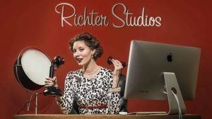 A woman sitting at a desk with a computer and a studio light. She is speaking into a vintage candlestick phone.