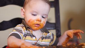 A baby sitting in a high chair and playing with his food. The baby food is smeared across his face and hands.