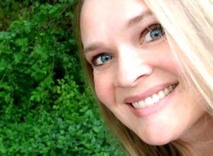 Emily Flegal, a woman with blond hair and blue eyes, taking a selfie in a forest.